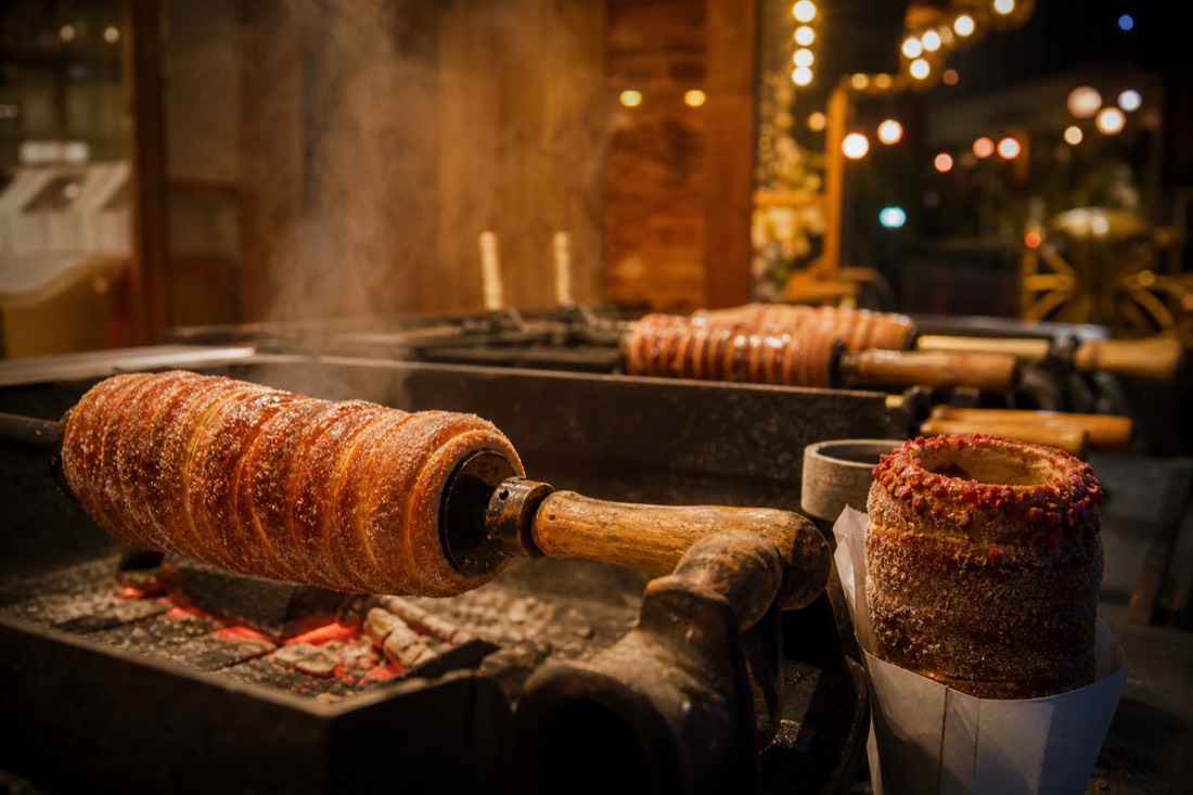 Close view of chimney cakes roasting on rotating wooden spits over glowing embers at a Budapest Christmas market stall, caramelized sugar glistening, one finished cake dusted with crushed red berries on a paper wrapper, steam curling up through strings of warm lights, night scene, no people in frame
