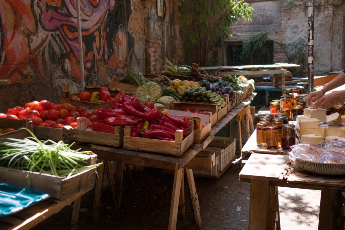Sunday morning farmers market inside a Budapest ruin bar courtyard, wooden crates of tomatoes and peppers on trestle tables, jars of honey and jam catching shafts of morning light through a broken roof, graffiti walls behind the produce, hands of a vendor arranging cheese, no faces visible