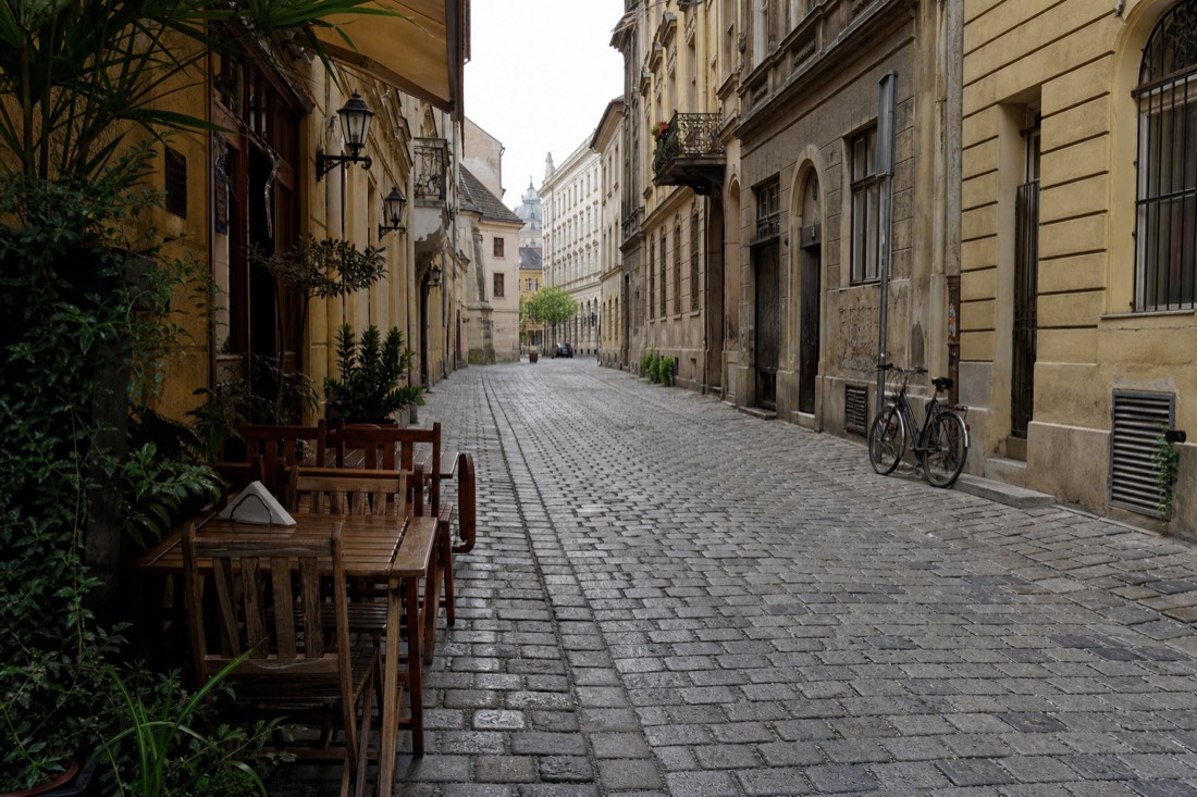 A cobbled Pest side street at mid morning, worn grey paving stones catching soft overcast light, ochre and cream facades with iron balconies, a small cafe terrace with empty wooden chairs and a folded napkin on one table, a bicycle leaning against a wall, quiet and unposed, no people prominent, no readable text or logos