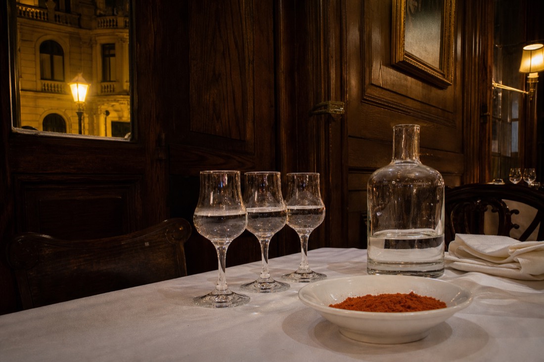 Three small stemmed pálinka glasses and a squat clear bottle on a white linen tablecloth in a warmly lit Budapest dining room, a shallow bowl of ground paprika and a folded napkin beside them, dark wood panelling and a shuttered window behind, evening lamplight, no people in frame and no readable text or logos