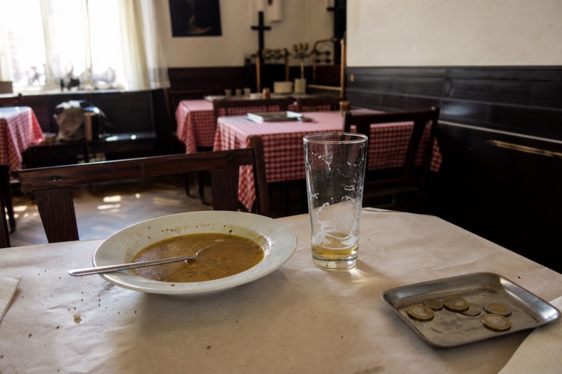 Interior of a small Budapest neighbourhood restaurant at lunchtime, a paper covered table with a bowl of soup, a half drunk glass of lager and a small metal bill tray holding forint coins, plain painted walls, worn parquet floor, soft daylight from a side window, quiet and unstaged, no people prominent, no readable text or logos