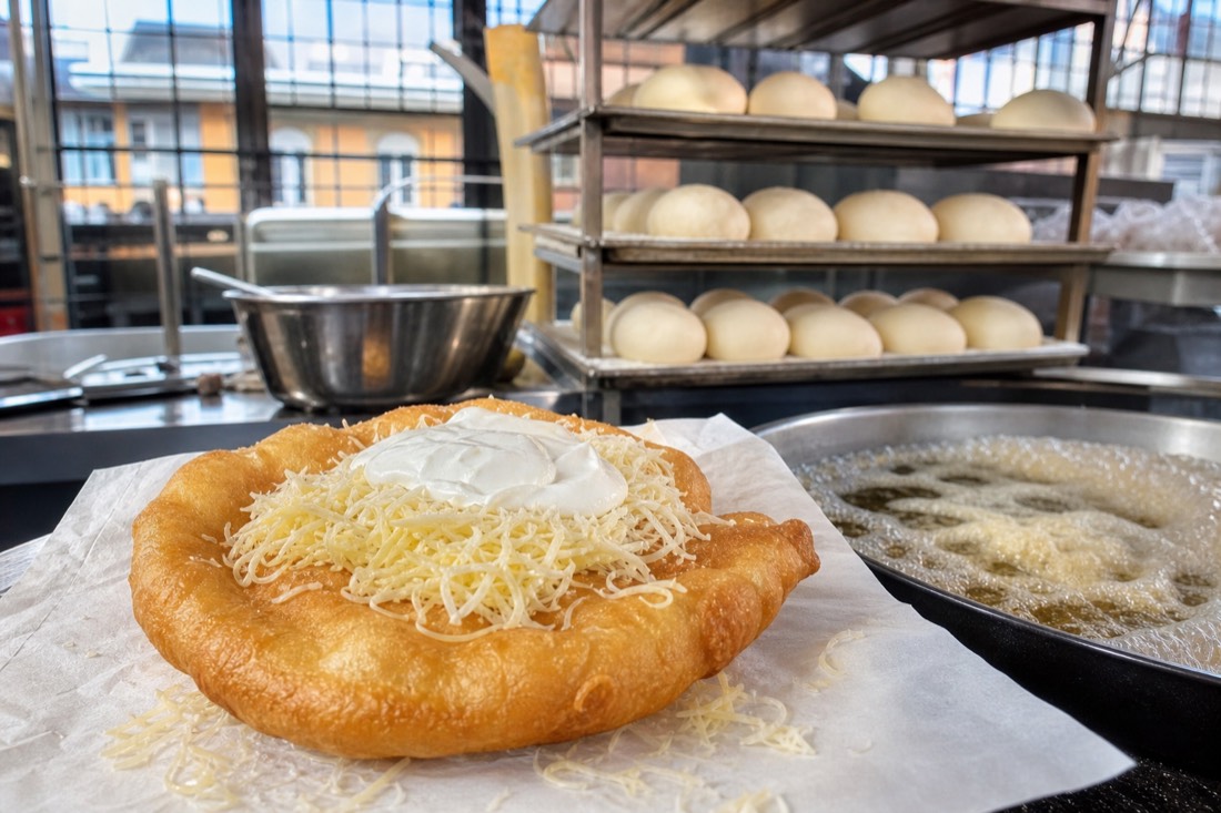 A fresh langos lifted from bubbling oil onto paper at a neighbourhood market counter in Budapest, blistered golden dough steaming under a scatter of grated cheese and a spoon of sour cream, stainless counter and stacked trays of pale dough behind, bright morning light through tall market hall windows, no people prominent, no readable signage
