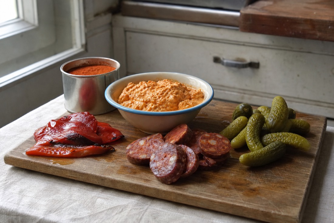 A wooden board on a linen cloth holding a bowl of paprika-red curd cheese spread, roast red peppers, sliced cured sausage and a heap of pickled cucumbers, a small tin of ground paprika beside it, absolutely no bread or pastry in the frame, soft daylight from a side window in a plain Budapest kitchen, no people and no readable text