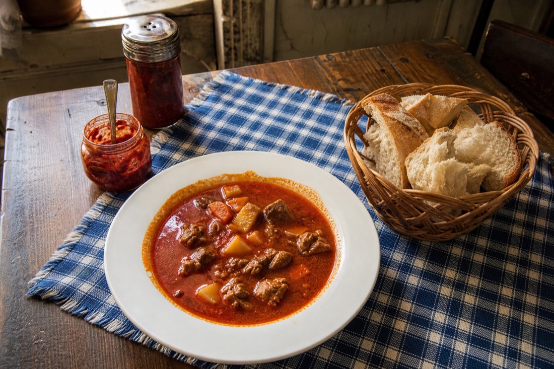 Overhead view of a Hungarian lunch table with a wide bowl of brick red gulyas soup, a basket of torn white bread, a small jar of coarse red hot pepper paste and a paprika shaker on a faded blue checked cloth, worn wooden table, soft daylight from a side window in an old Budapest dining room, no people, no readable text or labels
