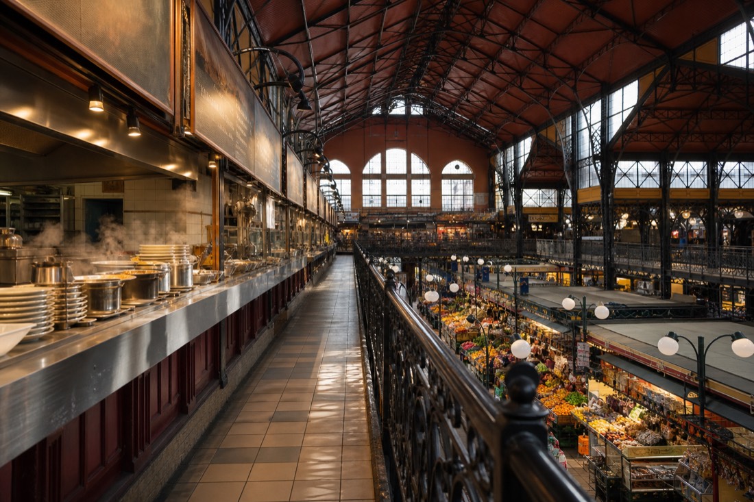 Upper gallery of Budapest's Great Market Hall in late morning, seen from the balcony rail: a row of small hot food counters with steaming pots and stacked plates, hand-lettered menu boards blurred beyond reading, warm light falling from the high steel and glass roof onto the produce aisles below, rich reds and browns, no people prominent, no readable text or logos