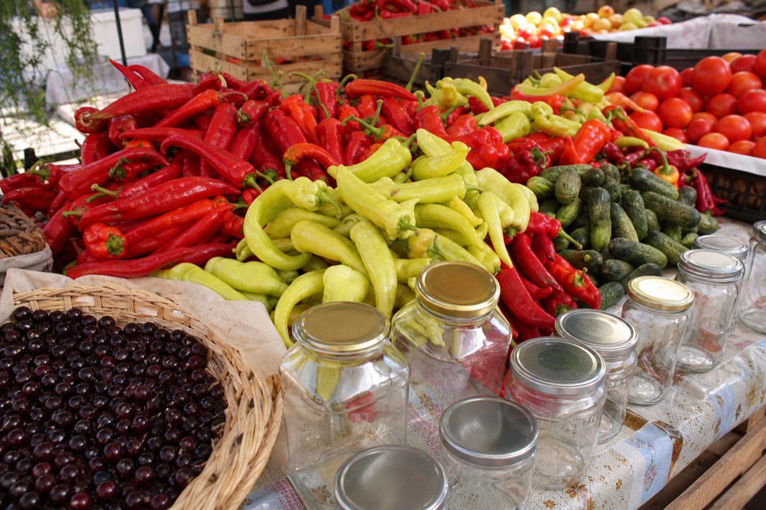 Late-season Hungarian market table loaded for preserving: heaps of long red and pale green pointed peppers, ripe tomatoes and small cucumbers beside empty glass jars with metal lids and a basket of dark sour cherries, plain wooden crates, bright natural daylight, warm reds and greens, no people prominent, no readable text or logos
