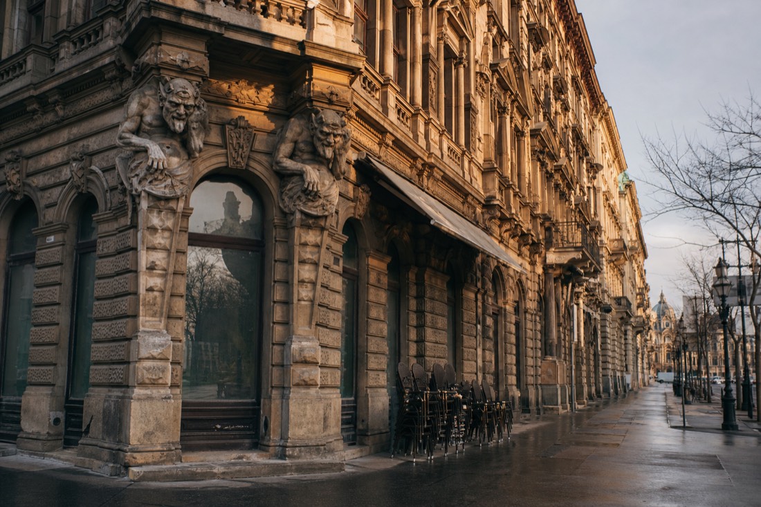 Early morning street view of an ornate 1890s palace facade on Budapest's Grand Boulevard, carved stone devil figures on the corner pilasters, damp pavement reflecting low golden sunlight, empty cafe terrace chairs stacked beside the entrance, warm ochre stonework against a pale winter sky, wide architectural view, no people prominent, no readable text or logos