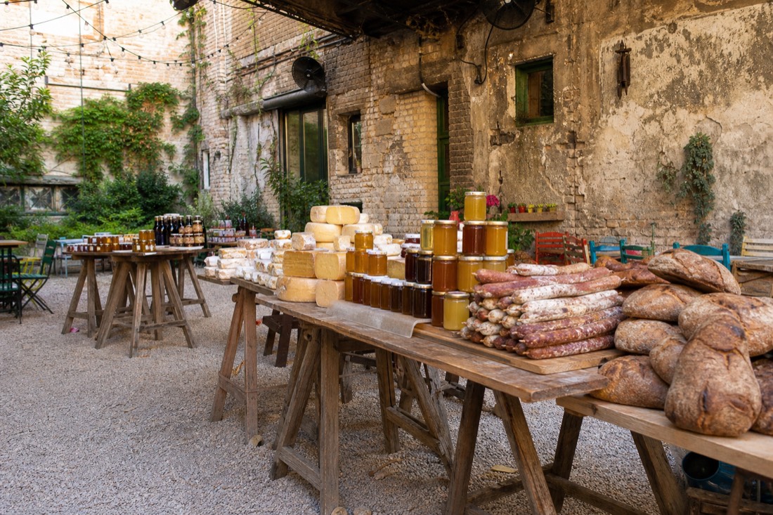 Sunday farmers market inside a Budapest ruin bar courtyard in bright daylight, trestle tables stacked with jars of honey, wheels of cheese, cured sausage and dark country bread, crumbling brick walls and unlit fairy light strings overhead, ivy and mismatched painted chairs pushed to the sides, soft diffused morning light, no people prominent, no readable text or logos