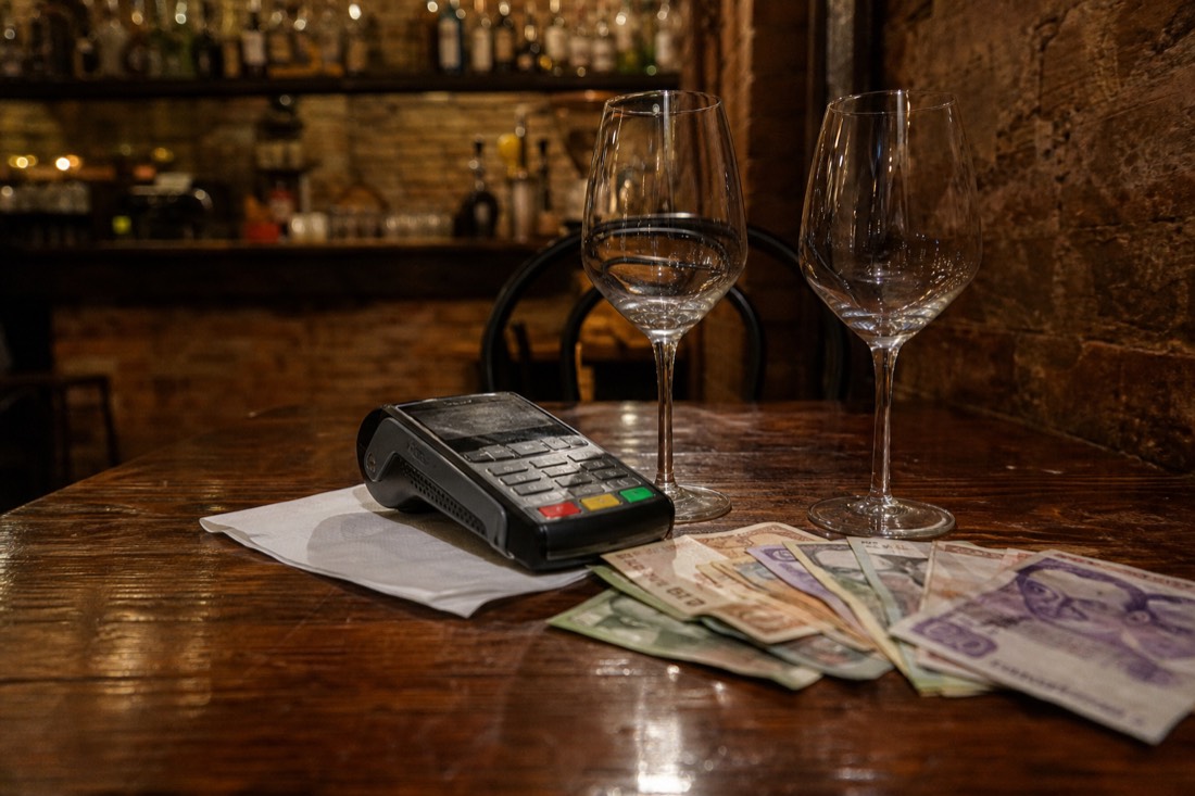 A handheld card payment terminal resting on a folded paper bill beside two empty wine glasses and a scatter of forint banknotes on a worn wooden table in a dim Budapest tavern, warm low lamplight, brick wall and shelves of bottles blurred in the background, no people in frame, no readable text or logos