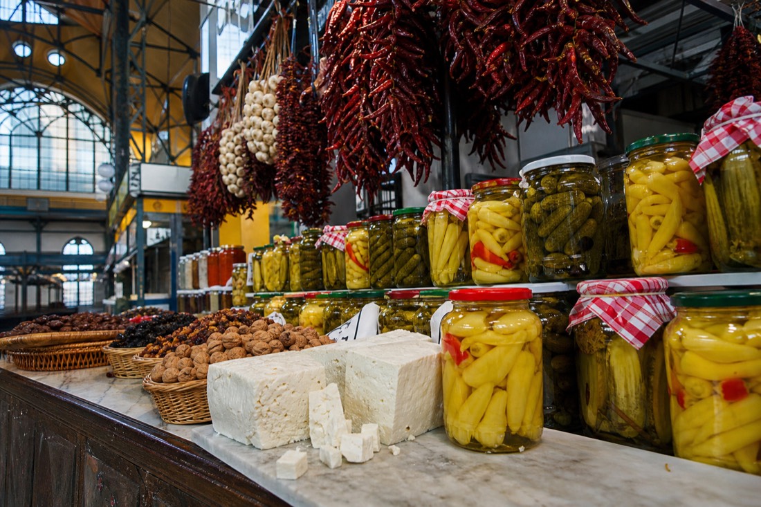 A Budapest market hall stall counter piled with glass jars of pickled cucumbers and yellow peppers, blocks of white sheep cheese on a marble slab, baskets of walnuts and dried fruit, strings of dried red paprika hanging above the counter, cool daylight from a high iron-framed roof window, no people in frame and no readable text or logos