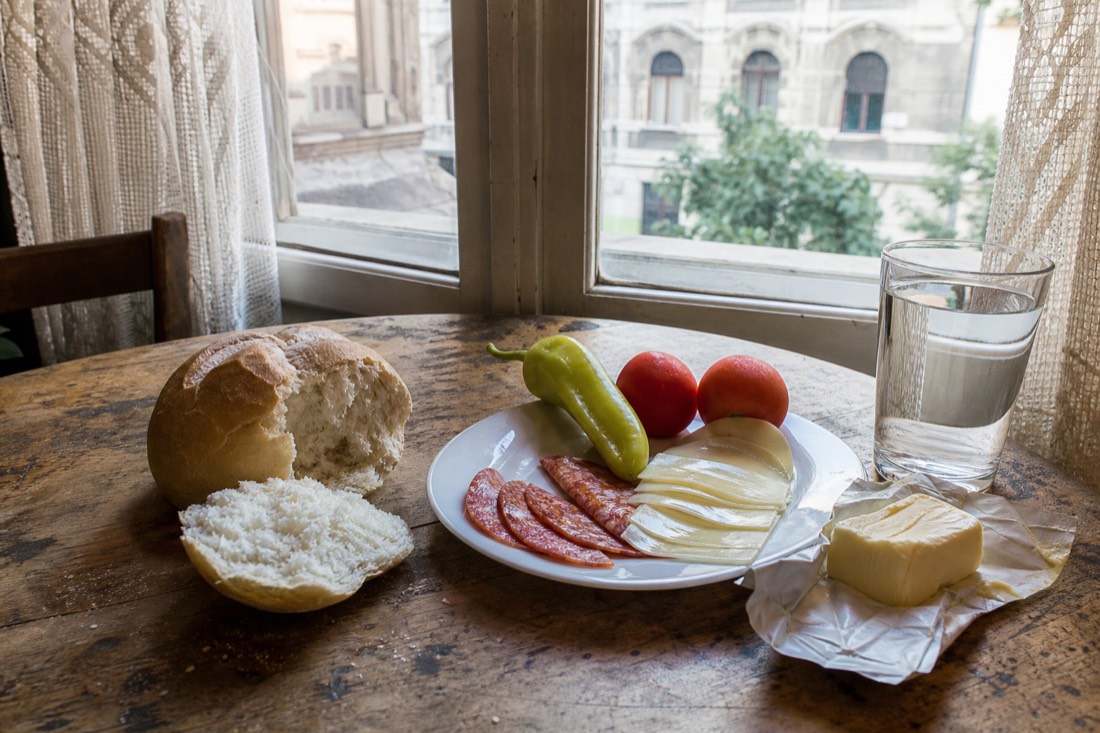 Simple Budapest breakfast laid on a worn wooden table by a window: fresh white bread rolls torn open, a slab of butter, thin slices of paprika salami and mild cheese on a plain white plate, a green pointed pepper and two tomatoes beside a glass of water, soft morning light through lace curtains, warm neutral tones, no people prominent, no readable text or logos