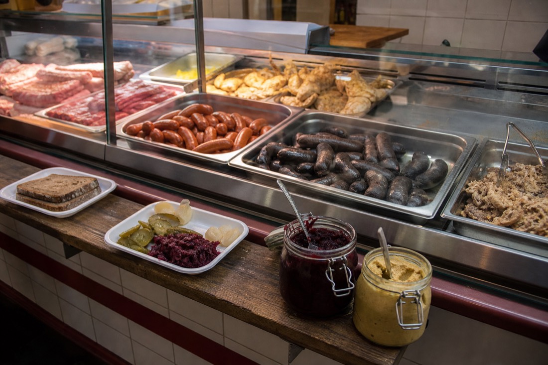Glass fronted butcher counter in a downtown Budapest shop, trays of paprika-red sausages and dark blood sausage under bright counter light, pots of mustard, sliced pickles and braised red cabbage beside a narrow standing ledge with paper plates and dark bread, tiled wall behind, warm practical lighting, no people prominent, no readable text or logos