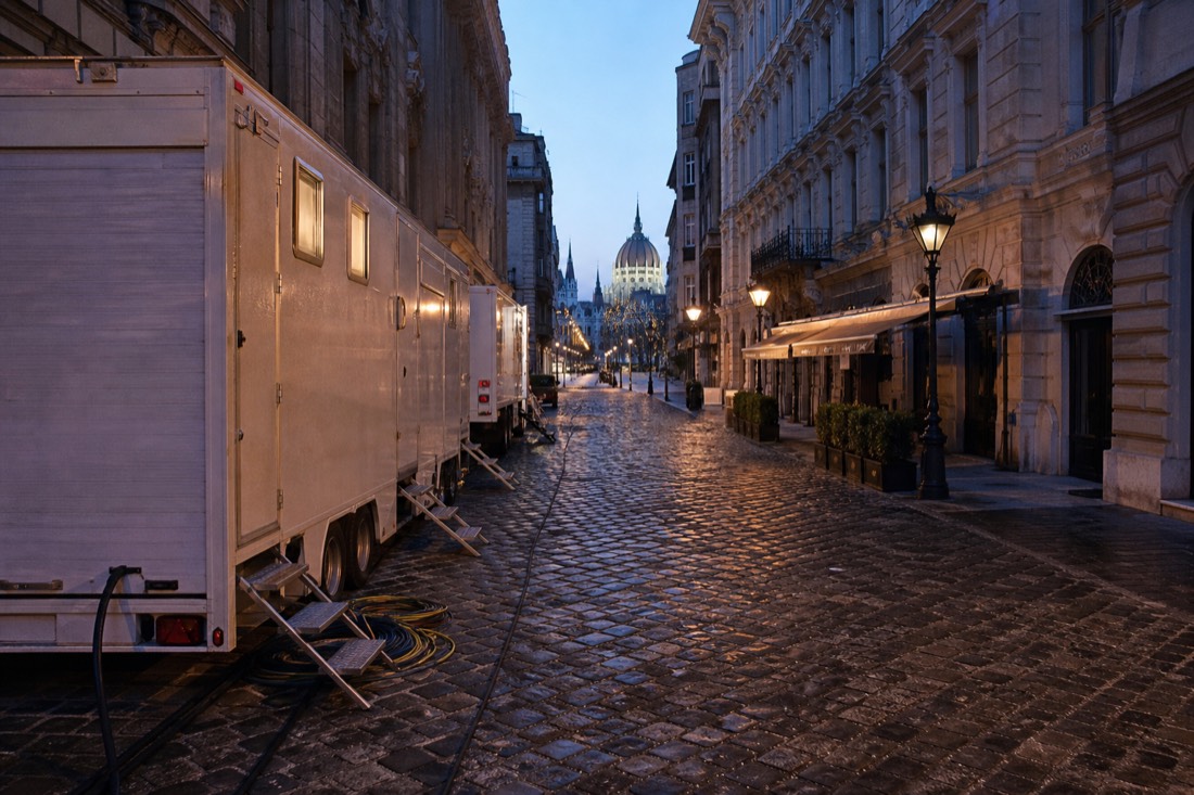 Cobbled central Budapest side street at dawn dressed for filming, empty production trailers and coiled cables along the kerb, nineteenth century stucco facades with shuttered cafe awnings, wet stones catching cold blue early light, long empty perspective down the street, no people prominent, no readable text or logos
