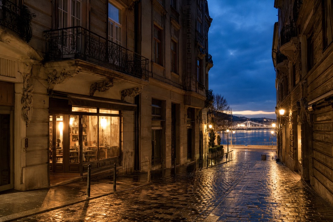 A quiet Pest street at dusk between old apartment buildings, warm light spilling from a ground floor cafe window onto wet cobbles, ornate iron balconies above, a narrow strip of the Danube and a lit bridge visible at the far end of the street, deep blue evening sky, no people prominent, no readable text or logos